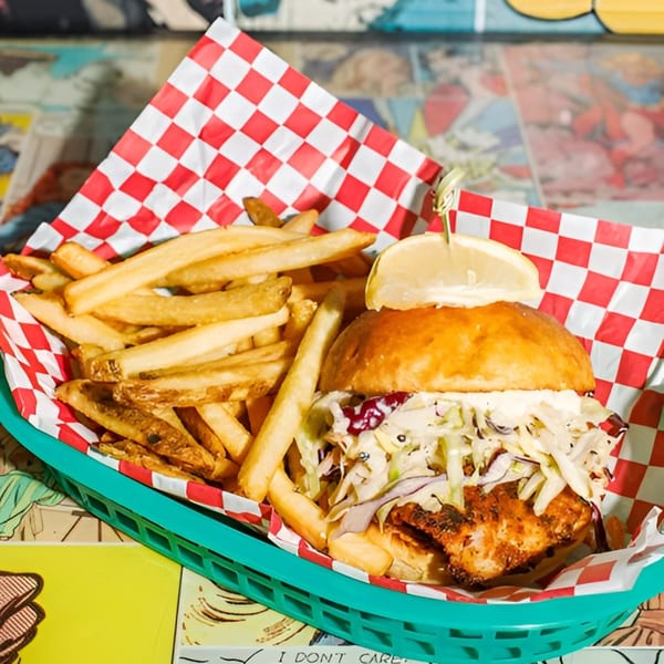 Crispy fish sandwich with coleslaw and a lemon wedge, served with fries in a green basket.