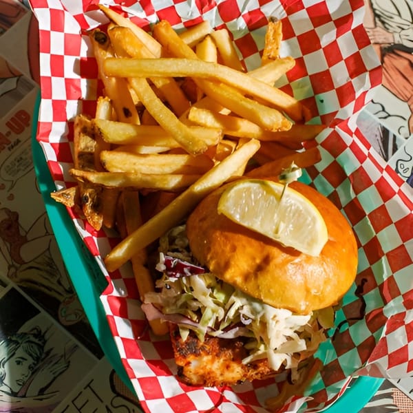 Fish sandwich with coleslaw and a lemon wedge, served with fries in a red and white checkered basket.