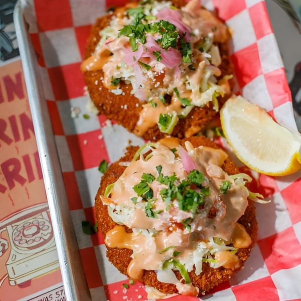 Two crispy crab cakes topped with slaw, pink pickled onions, sauce, and cilantro, with a lemon wedge.