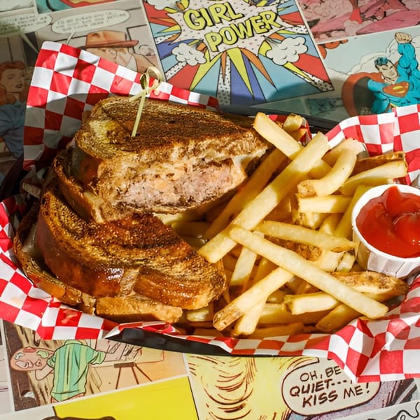 A grilled sandwich and fries with ketchup in a basket on a comic book themed table.