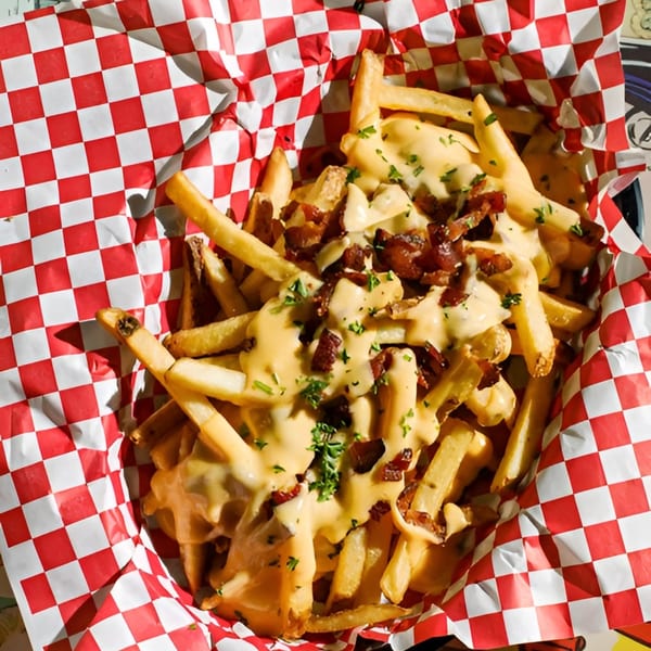 Golden french fries topped with melted cheese, crispy bacon bits, and fresh parsley in a red and white checkered basket.