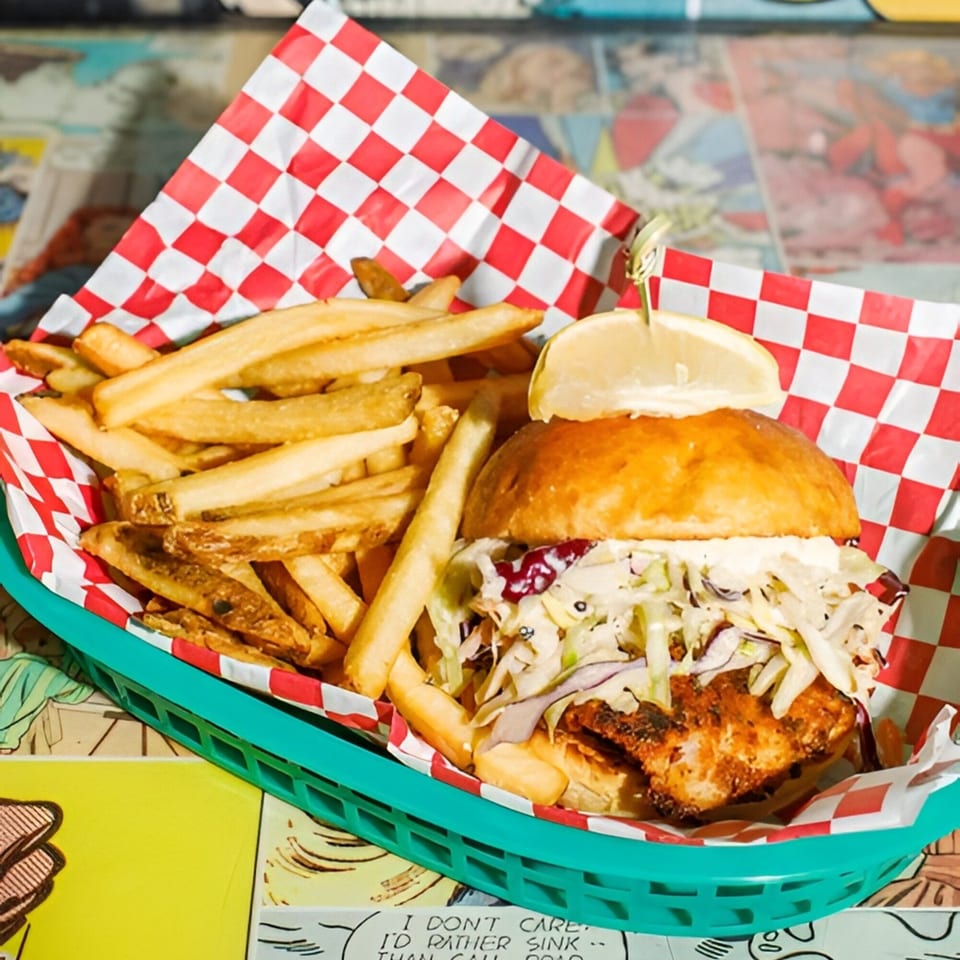 Crispy fish sandwich with coleslaw and fries in a green basket with red checkered paper.