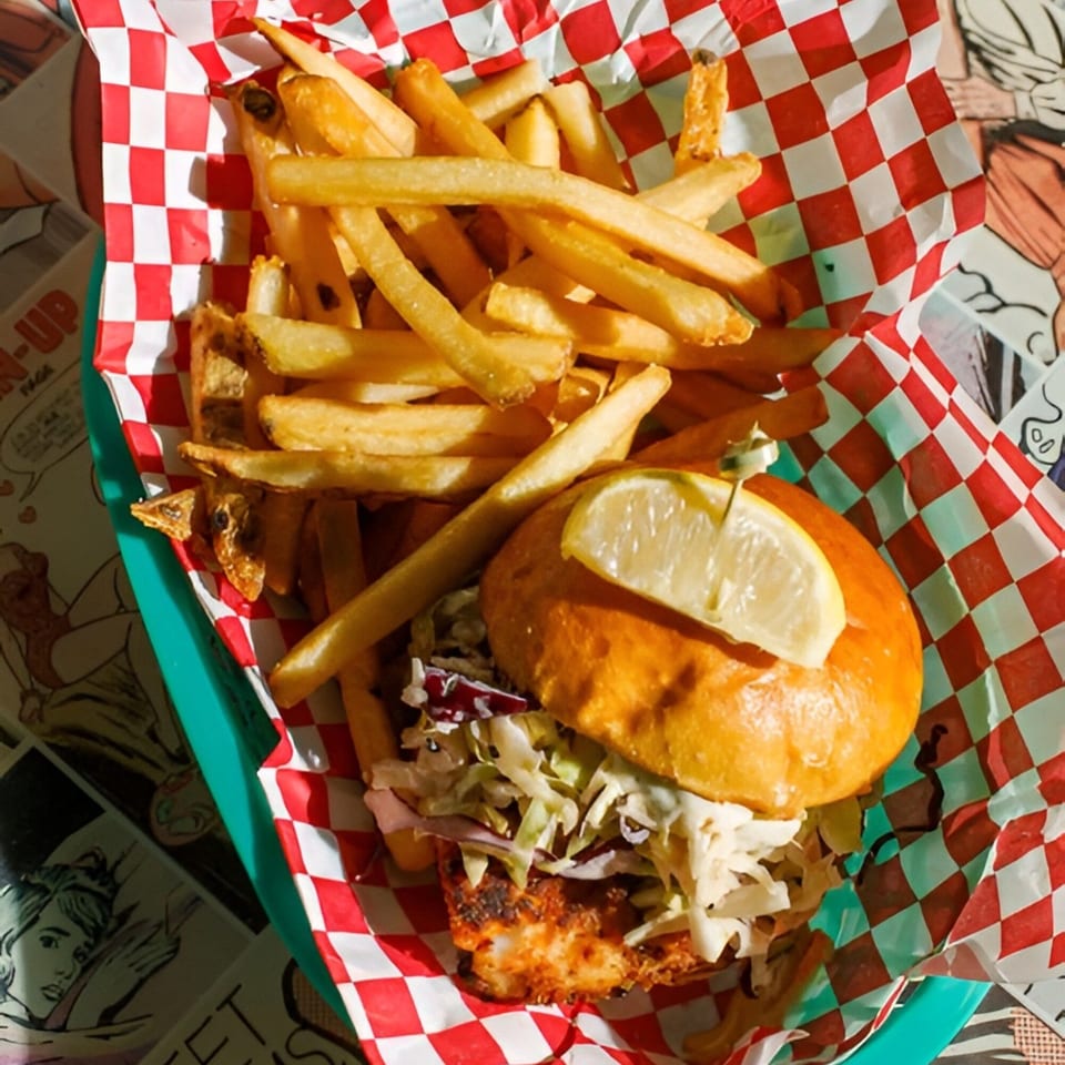 A fish sandwich with coleslaw and a lemon wedge, served with fries in a red and white checkered basket.