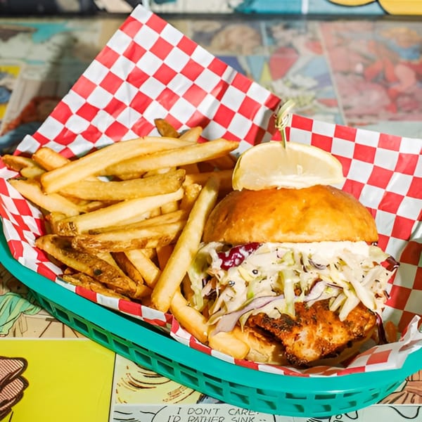 A fish sandwich with coleslaw and a lemon wedge, served with fries in a green basket.