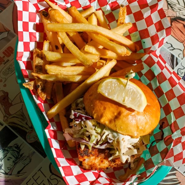 Fish sandwich with coleslaw and a lemon wedge, served with fries in a red and white checkered basket.