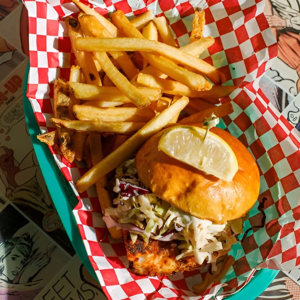Fish sandwich with coleslaw and a lemon wedge, served with fries in a red and white checkered basket.