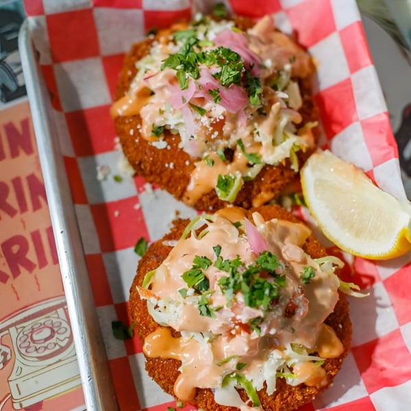 Two crispy crab cakes topped with slaw, pink pickled onions, sauce, and cilantro, with a lemon wedge.