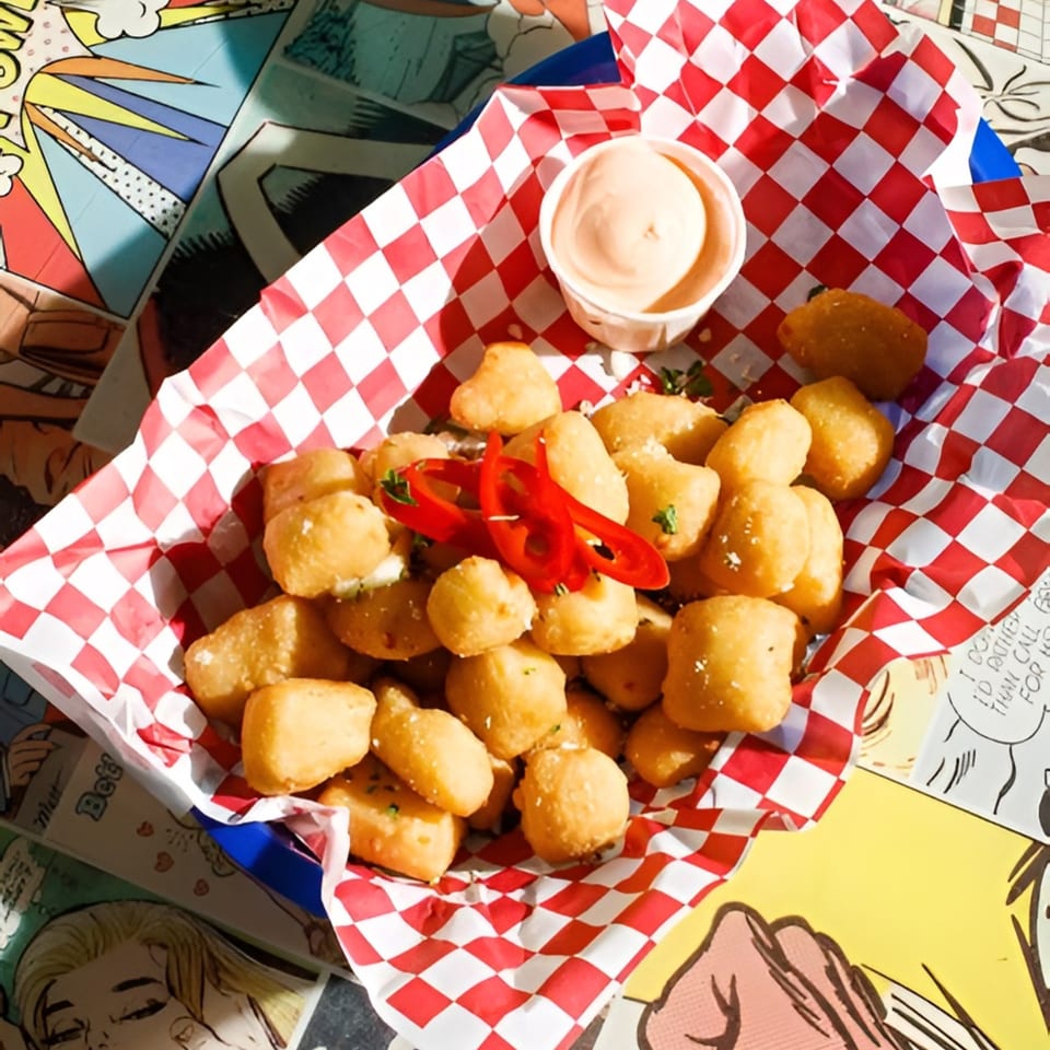 Golden fried cheese curds with red chili peppers and a side of dipping sauce in a red checkered basket.